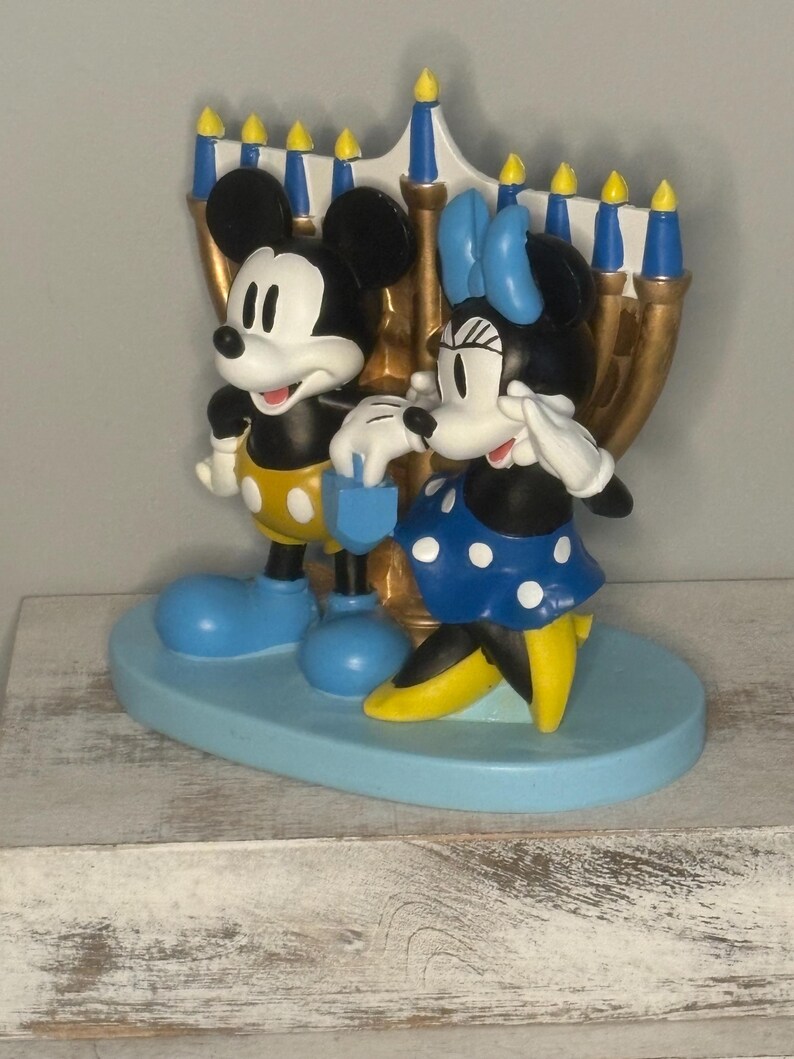 May include: A decorative figurine featuring Mickey and Minnie Mouse standing in front of a gold-colored menorah. Mickey wears yellow shorts and blue shoes, while Minnie dons a blue polka-dot dress and yellow shoes. The base is light blue.