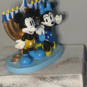 May include: A decorative figurine featuring Mickey Mouse and Minnie Mouse standing in front of a menorah. The characters are in their classic colors, with Mickey in yellow shorts and Minnie in a blue dress. The menorah is gold with blue and yellow accents.