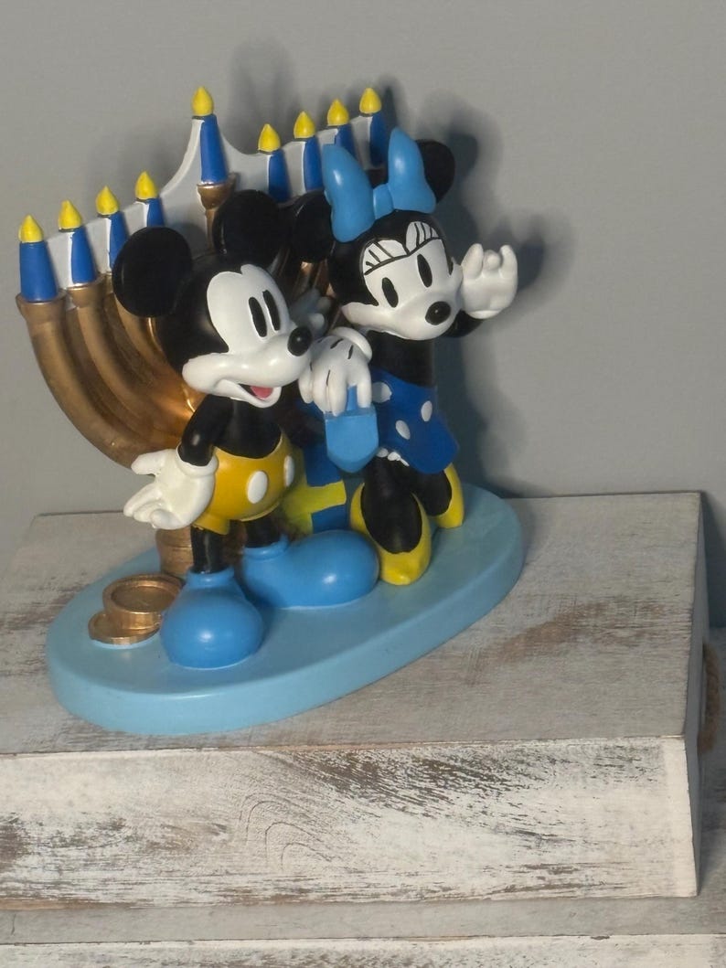 May include: A decorative figurine featuring Mickey Mouse and Minnie Mouse standing in front of a menorah. The characters are in their classic colors, with Mickey in yellow shorts and Minnie in a blue dress. The menorah is gold with blue and yellow accents.