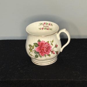 May include: A white ceramic mug with a large pink rose and green leaves. The word "June" is printed above the rim, surrounded by small pink flowers. The mug has a gold rim and handle.