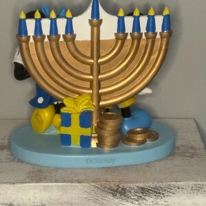 May include: A gold-colored menorah with blue and yellow candles, featuring Mickey Mouse figures. The base is light blue, with a yellow and blue gift box and gold coins. The Disney logo is visible on the base.