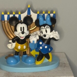 May include: A decorative figurine featuring Mickey and Minnie Mouse in front of a gold menorah. Mickey wears yellow shorts and blue shoes, while Minnie dons a blue polka-dot skirt and bow. The base is light blue.