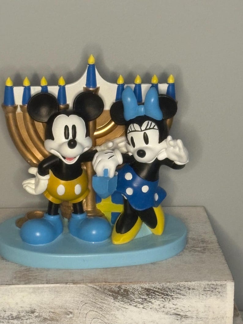 May include: A decorative figurine featuring Mickey and Minnie Mouse in front of a gold menorah. Mickey wears yellow shorts and blue shoes, while Minnie dons a blue polka-dot skirt and bow. The base is light blue.