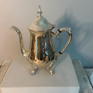 May include: A polished silver-toned metal teapot featuring a tall, pointed lid, a curved spout, and an ornate handle with two black circular accents. The teapot stands on three decorative feet and has a reflective surface showing surrounding light.