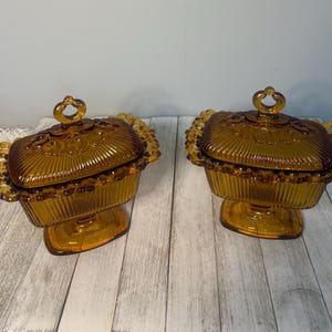 May include: Two amber-colored glass dishes with matching lids. Each rectangular dish features vertical ribbed sides, a scalloped edge, and a central loop handle on the lid. The glass has a translucent, warm brown tone with a glossy finish.