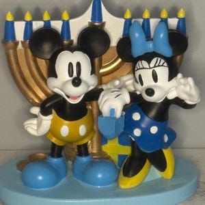 May include: A decorative figurine featuring Mickey and Minnie Mouse standing in front of a gold menorah. Mickey wears yellow shorts and blue shoes. Minnie wears a blue polka-dot dress and bow. The base is light blue.