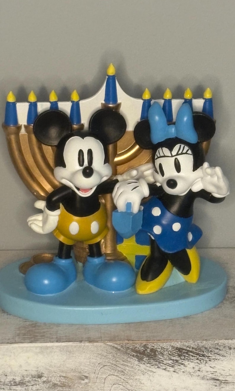 May include: A decorative figurine featuring Mickey and Minnie Mouse standing in front of a gold menorah. Mickey wears yellow shorts and blue shoes. Minnie wears a blue polka-dot dress and bow. The base is light blue.
