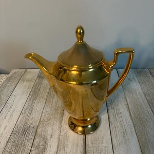 May include: A gold-colored teapot with a lid and a handle. The teapot has a spout and sits on a small, round base. The surface of the teapot is shiny and reflective, and the overall design is elegant and classic.