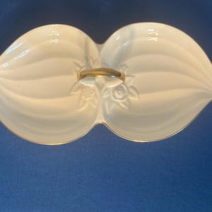 May include: A white, two-sectioned serving dish with a gold handle and trim. The dish has a leaf-like shape with embossed floral details. The background is a solid blue.