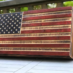 Op de afbeelding: Een Amerikaanse houten vlag met een verweerde afwerking. De vlag is gemaakt van afzonderlijke houten planken en heeft een donker rood, wit en blauw kleurenschema. De vlag is gemonteerd op een houten frame.