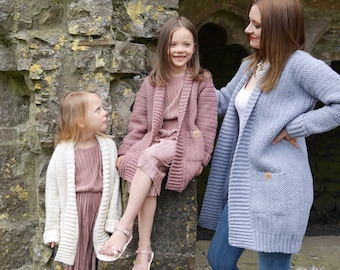 May include: Three girls wearing cardigans in different colors. The girl in the middle is wearing a pink cardigan, the girl on the left is wearing a white cardigan, and the girl on the right is wearing a blue cardigan.