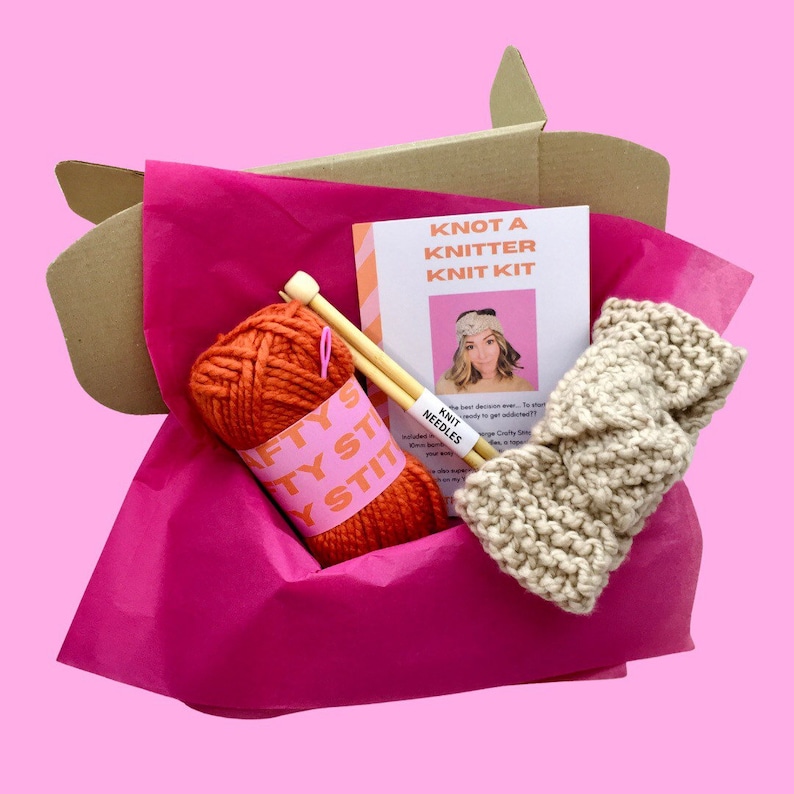 May include: A brown cardboard box filled with pink tissue paper. The box contains a kit for knitting a headband. The kit includes orange yarn, wooden knitting needles, and a beige knitted headband. The box also includes a printed instruction sheet with the title "Knot a Knitter Knit Kit".