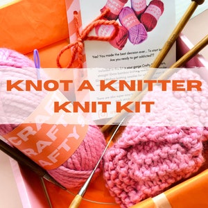 Könnte beinhalten: Ein rosa-oranges Strickset mit dem Text "KNOT A KNITTER KNIT KIT". Das Set enthält rosa Garn, Stricknadeln und ein gestricktes Muster. Das Set befindet sich in einer rosa Box mit orangefarbenen Akzenten.