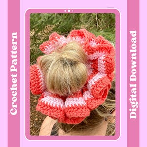 May include: A crochet hair scrunchie in pink and coral colors. The scrunchie is made with a textured stitch and has a ruffled edge. The scrunchie is being worn by a person with blonde hair.