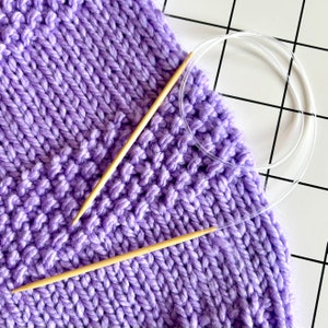 Può includere: Tessuto a maglia viola con due ferri da maglia in legno e un ago circolare in plastica trasparente.