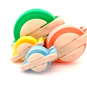 May include: Four colourful yarn ball winders, green, yellow, blue, and pink, are stacked on top of each other. The winders are made of plastic and have a white centre.