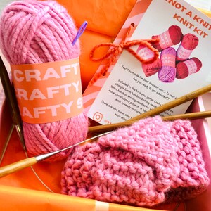 May include: A pink knitting kit containing a ball of yarn, two wooden knitting needles, and a partially knitted pink scarf. The kit includes instructions and is labelled "Not a Knitter Knit Kit".