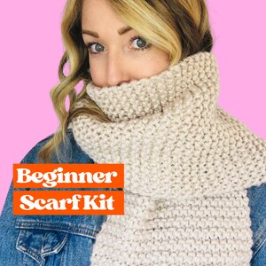 May include: A woman wearing a beige knitted scarf. The scarf is thick and textured. The text "Beginner Scarf Kit" is displayed in orange letters on a white background.