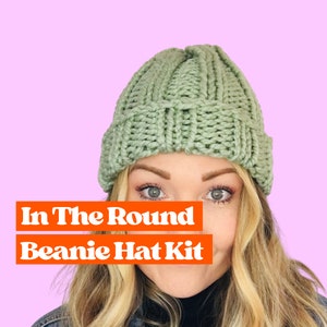 May include: A woman wearing a sage green knitted beanie hat. The hat is made with a chunky yarn and has a ribbed texture. The text "In The Round Beanie Hat Kit" is displayed in orange letters.