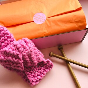 May include: A pink knitted headband with a pink and white label that says "That Crafty Stitch" on a pink and orange background. The headband is next to two wooden knitting needles.