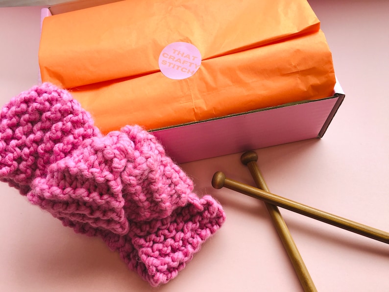 May include: A pink knitted headband with a pink and white label that says "That Crafty Stitch" on a pink and orange background. The headband is next to two wooden knitting needles.
