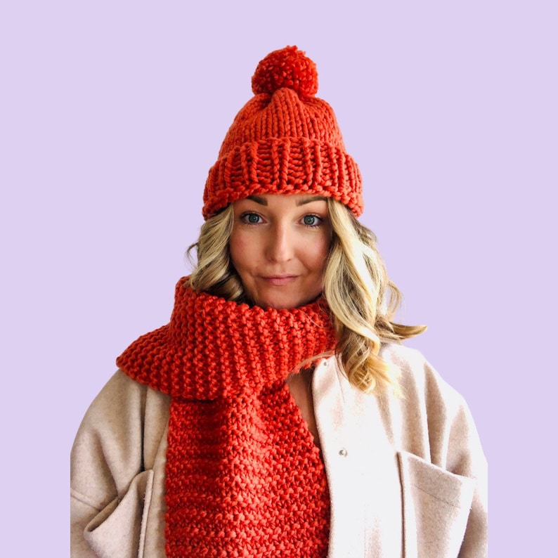 May include: A bright orange knitted hat with a pom pom on top and a matching scarf. The hat and scarf are made of a chunky yarn.