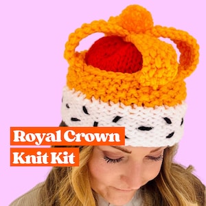 Op de afbeelding: Een gebreide muts in de vorm van een kroon met een rode pompon bovenop. De kroon is oranje en wit met zwarte accenten. De muts wordt gedragen door een persoon met blond haar. De tekst "Royal Crown Knit Kit" wordt onder de muts weergegeven.
