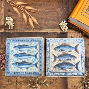 May include: Two square ceramic tiles with blue and white floral designs and three blue fish on each tile.
