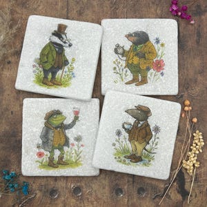 Puede incluir: Juego de cuatro posavasos cuadrados con ilustraciones caprichosas de animales antropomórficos. Cada posavasos representa un personaje diferente: un tejón, un topo, una rana y una rata, todos vestidos con atuendos vintage y sobre un fondo floral.