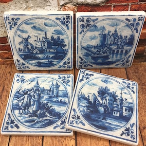 Könnte beinhalten: Vier quadratische Keramikfliesen mit blau-weißen Delftware-Designs. Jede Fliese zeigt eine andere Szene, darunter eine Windmühle, ein Schiff, ein Schloss und eine Brücke. Die Fliesen sind perfekt, um jedem Zuhause einen Hauch von holländischem Charme zu verleihen.