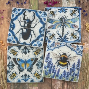 Pode incluir: Quatro bases de cerâmica quadradas com desenhos de insetos. Cada base apresenta um inseto diferente: um besouro, uma libélula, uma borboleta e um zangão. As bases têm um fundo branco com bordas florais azuis.