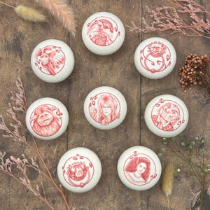 May include: Eight round white ceramic knobs featuring detailed red line art illustrations of various characters and motifs. Each knob displays a unique circular design, including portraits of people and creatures, arranged in a circular pattern.