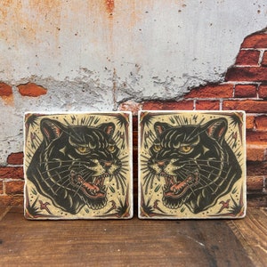 Könnte beinhalten: Zwei quadratische Untersetzer aus Keramik mit einem schwarzen Pantherkopf-Design. Der Panther hat einen grimmigen Ausdruck mit scharfen Zähnen und gelben Augen. Der Hintergrund ist hellbeige mit einem dunkelbraunen Rand und einem Muster aus schwarzen Blitzen.