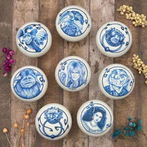 May include: Eight white ceramic cabinet knobs with blue illustrations. The knobs depict various character portraits and designs, including a door knocker and a woman's portrait. The knobs are arranged on a wooden surface.