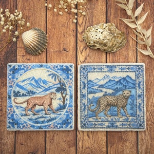 May include: Two square ceramic coasters with blue and white floral borders. Each coaster features a different leopard print design with a blue and white landscape background.