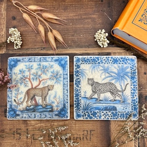 Könnte beinhalten: Zwei quadratische Keramikuntersetzer mit blauen und weißen Blumendesigns. Jeder Untersetzer zeigt ein schwarz-weißes Leopardenmustertier in einer anderen Pose. Ein Untersetzer zeigt den Leoparden in einer Dschungelumgebung, während der andere den Leoparden in einer Gartenumgebung zeigt.