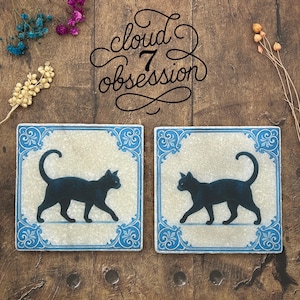 May include: Two square coasters with a black cat silhouette on a cream background, framed by a blue and white decorative border. The text "cloud 7 obsession" is written in a cursive font above the coasters.