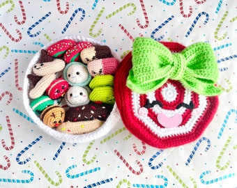Op de afbeelding: Een witte gehaakte mand gevuld met verschillende gehaakte etenswaren, waaronder een donut, een hotdog, een macaron en een watermeloenplak. De mand staat naast een rood-witte gehaakte snoepstok met een groene strik.