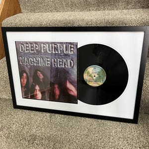 May include: Framed Deep Purple "Machine Head" album. The album cover features a blurred image of the band members. The vinyl record is displayed next to the album cover in a white mat and black frame. The album is displayed on a carpeted surface.