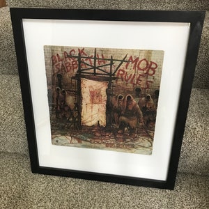 May include: Framed print of the Black Sabbath album "Mob Rules." The artwork features a dark, detailed illustration with figures in hooded cloaks, a brick wall, and the album title in red graffiti-style lettering. The frame is black.