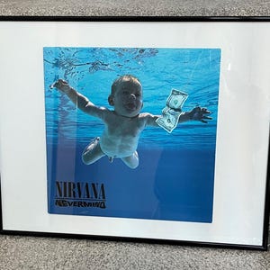 May include: A framed poster of the Nirvana album "Nevermind". The poster features a black and white photo of a baby swimming underwater, reaching for a dollar bill. The album title is printed in black text at the bottom of the poster.