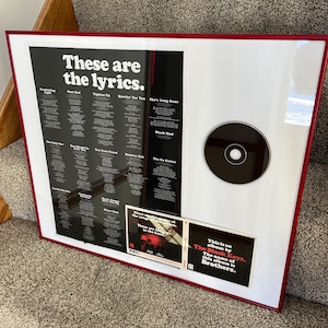 May include: Framed artwork with a red border, featuring a black and white print of song lyrics titled "These are the lyrics." Includes a CD and two small inserts. The album title is "Brothers" by The Black Keys.