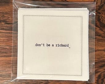 Puede incluir: Un conjunto de posavasos cuadrados de color blanquecino en una funda de plástico transparente. Cada posavasos tiene un borde punteado y la frase "don't be a richard" impresa en morado oscuro.