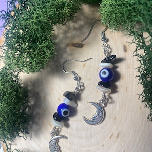 Könnte beinhalten: Ein Paar silberne Ohrringe mit blauen bösen Augen-Anhängern, schwarzen Perlen, weißen Muschelperlen und Halbmond-Anhängern.