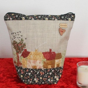 Peut inclure: Un sac fourre-tout beige avec un motif de paysage rural, représentant des maisons et des arbres, est présenté sur une surface rouge. Le sac a une base et une bordure à motifs floraux noirs. Une boîte en bois et une bougie dans un verre sont également sur la surface.