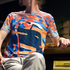 May include: A short-sleeved t-shirt with a colorful abstract design featuring orange, blue, and yellow shapes. The shirt has a crew neck and a graphic print with the word "crossroad". The person is wearing light gray pants.