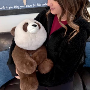 May include: A plush panda bear with a white face and chest, brown ears, paws, and body. The panda has a black nose and a smiling expression. The stuffed animal is being held.