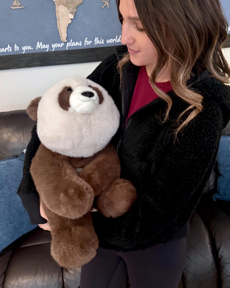 May include: A plush panda bear with a white face and chest, brown ears, paws, and body. The panda has a black nose and a smiling expression. The stuffed animal is being held.