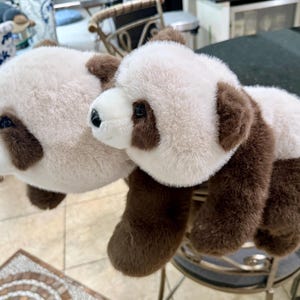 May include: Two plush panda bears with white and brown fur. The pandas have black eyes and noses. The bears are lying on a black surface. The panda bears are a cute and cuddly toy.