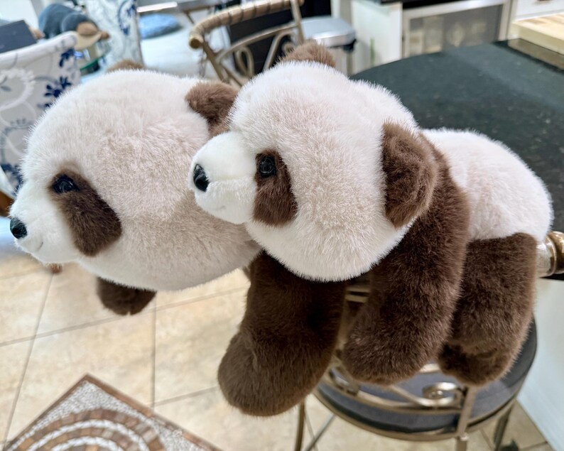 May include: Two plush panda bears with white and brown fur. The pandas have black eyes and noses. The bears are lying on a black surface. The panda bears are a cute and cuddly toy.